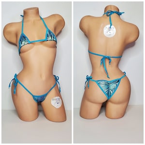 Puede incluir: Un conjunto de bikini turquesa con un top halter y una parte inferior atrevida. El bikini está hecho de una tela brillante e iridiscente con un patrón azul y verde. El bikini se ata en el cuello y la espalda, y la parte inferior tiene lazos en los lados.