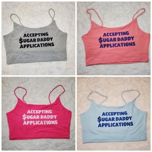 May include: Four crop tops in different colors: gray, pink, hot pink, and light blue. Each top has the same text printed on it: "ACCEPTING $UGAR DADDY APPLICATIONS".