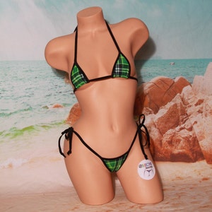 Puede incluir: Bikini de cuadros verdes y negros con ribetes negros. La parte superior del bikini es de estilo triangular y la parte inferior es de estilo atrevido con lazos a los lados. El bikini está en un maniquí sobre un fondo de playa.