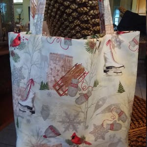 Puede incluir: Una bolsa de lona blanca con un estampado de temática invernal que presenta cardenales, copos de nieve, patines de hielo, guantes y un trineo. La bolsa tiene dos asas largas.