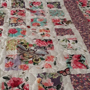 May include: A white quilt with a floral pattern. The quilt is made up of squares of different floral fabrics. The quilt has a bee with the text "Bee a Blessing" embroidered on it. The quilt is decorated with small white dots and has a purple and white floral border.