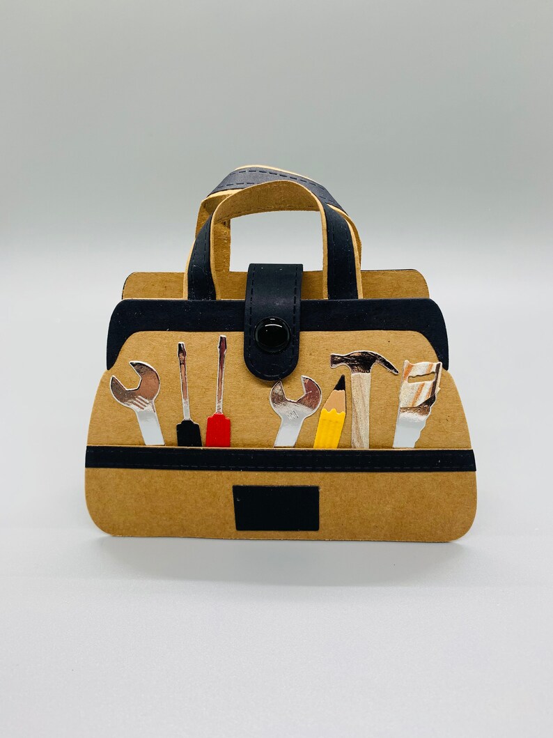 K&ouml;nnte beinhalten: Eine braune Papiertasche f&uuml;r Werkzeug mit einem schwarzen Griff und einer schwarzen Klappe. Die Tasche ist gef&uuml;llt mit verschiedenen Werkzeugen, darunter Schraubenschl&uuml;ssel, Schraubendreher, ein Hammer, ein Bleistift und eine S&auml;ge.
