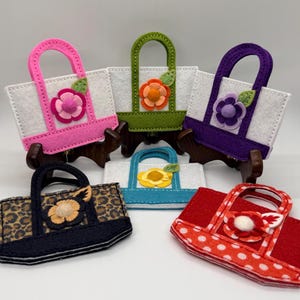 May include: A collection of felt tote bags in various colors and patterns. The bags feature floral embellishments and contrasting handles. Designs include pink, green, purple, blue, leopard print, and red with white polka dots. Each bag has a unique flower design.