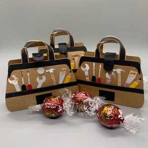 K&ouml;nnte beinhalten: Drei braune Werkzeugtaschen-Geschenkboxen aus Papier mit schwarzen Griffen und schwarzen Druckkn&ouml;pfen. Jede Box ist mit wei&szlig;en Werkzeugsilhouetten verziert. Drei rote Schokoladenbonbons liegen vor den Boxen.