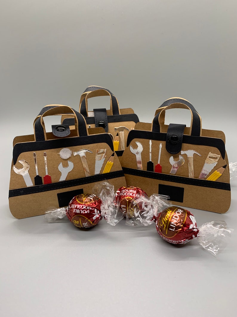 K&ouml;nnte beinhalten: Drei braune Werkzeugtaschen-Geschenkboxen aus Papier mit schwarzen Griffen und schwarzen Druckkn&ouml;pfen. Jede Box ist mit wei&szlig;en Werkzeugsilhouetten verziert. Drei rote Schokoladenbonbons liegen vor den Boxen.