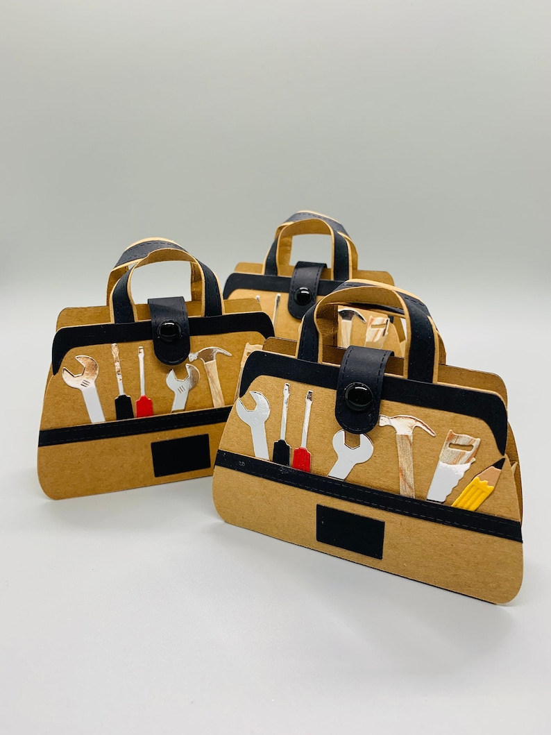K&ouml;nnte beinhalten: Drei braune Werkzeugtaschen-Geschenkboxen aus Papier mit schwarzen Griffen und schwarzen Akzenten. Jede Box hat eine Vielzahl von Werkzeugen auf der Vorderseite gedruckt, darunter Schraubenschl&uuml;ssel, Schraubendreher, einen Hammer, eine S&auml;ge und einen Bleistift.