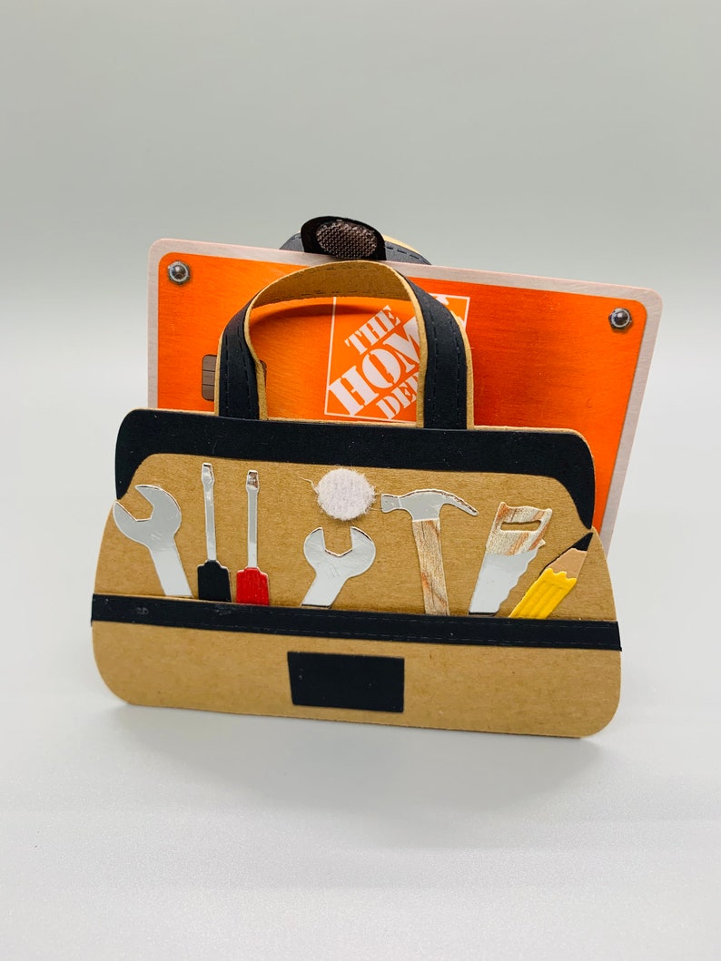K&ouml;nnte beinhalten: Eine braune Papiert&uuml;te in Form einer Werkzeugtasche mit einem schwarzen Griff und einem schwarzen Besatz. Die Tasche enth&auml;lt verschiedene Werkzeuge, darunter einen Hammer, einen Schraubenschl&uuml;ssel, einen Schraubendreher, eine S&auml;ge und einen Bleistift. Die Tasche liegt auf einer Geschenkkarte f&uuml;r The Home Depot.
