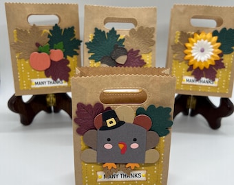 May include: Four brown paper gift bags with cut-out designs for Thanksgiving. Each bag has a different design, including a turkey, pumpkins, acorns, and a sunflower. The bags have the text "MANY THANKS" on them.
