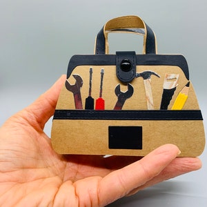 K&ouml;nnte beinhalten: Eine braune Papiert&uuml;te in Form einer Werkzeugtasche mit einem schwarzen Griff und einem schwarzen Knopfverschluss. Die Tasche ist gef&uuml;llt mit verschiedenen Werkzeugen, darunter ein Hammer, ein Schraubenschl&uuml;ssel, ein Schraubendreher und ein Bleistift.