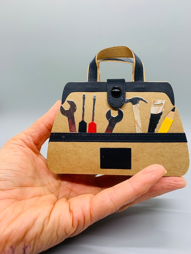 K&ouml;nnte beinhalten: Eine braune Papiert&uuml;te in Form einer Werkzeugtasche mit einem schwarzen Griff und einem schwarzen Knopfverschluss. Die Tasche ist gef&uuml;llt mit verschiedenen Werkzeugen, darunter ein Hammer, ein Schraubenschl&uuml;ssel, ein Schraubendreher und ein Bleistift.