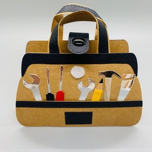 K&ouml;nnte beinhalten: Eine braune Papiertasche f&uuml;r Werkzeuge mit einem schwarzen Griff und einem schwarzen Knopfverschluss. Die Tasche ist gef&uuml;llt mit verschiedenen Werkzeugen, darunter ein Schraubenschl&uuml;ssel, ein Schraubendreher, ein Hammer, ein Bleistift und eine S&auml;ge.