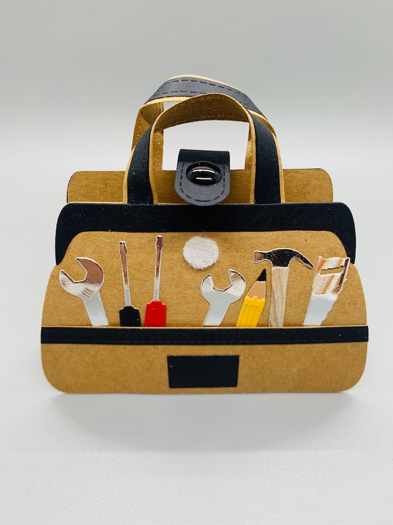 K&ouml;nnte beinhalten: Eine braune Papiertasche f&uuml;r Werkzeuge mit einem schwarzen Griff und einem schwarzen Knopfverschluss. Die Tasche ist gef&uuml;llt mit verschiedenen Werkzeugen, darunter ein Schraubenschl&uuml;ssel, ein Schraubendreher, ein Hammer, ein Bleistift und eine S&auml;ge.