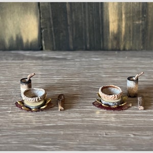 May include: Two miniature wooden bowls with a brown and white design, each sitting on a wooden base with gold accents. A small wooden stick is next to each bowl.