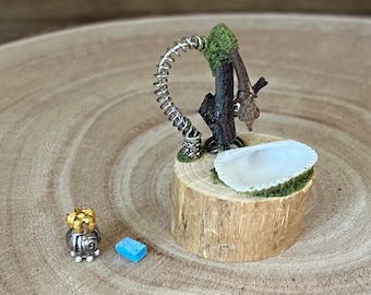 May include: Miniature fairy garden scene featuring a tree trunk with a silver faucet, a seashell-shaped tub, and a small gift. A blue rectangular soap and a small silver and gold gift are also present. The scene is set on a wooden slice.