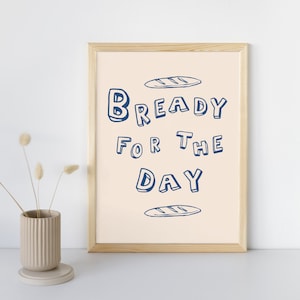 May include: A framed print with a light beige background and blue text that reads "Bready for the Day". The text is stylized with a bread-like outline. There are two small drawings of bread loaves above and below the text.