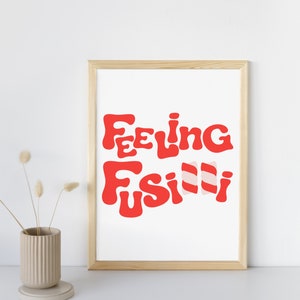 May include: A framed print with the text "Feeling Fusilli" in red and pink lettering. The print is in a light wood frame and is sitting on a white surface.