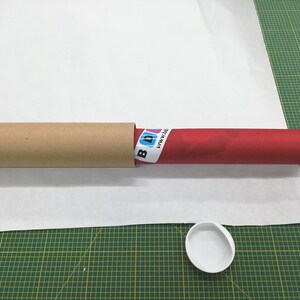 May include: A red poster tube with a white label that says "www.pixi.de" is inside a brown cardboard tube. A white plastic lid is on a green cutting mat.