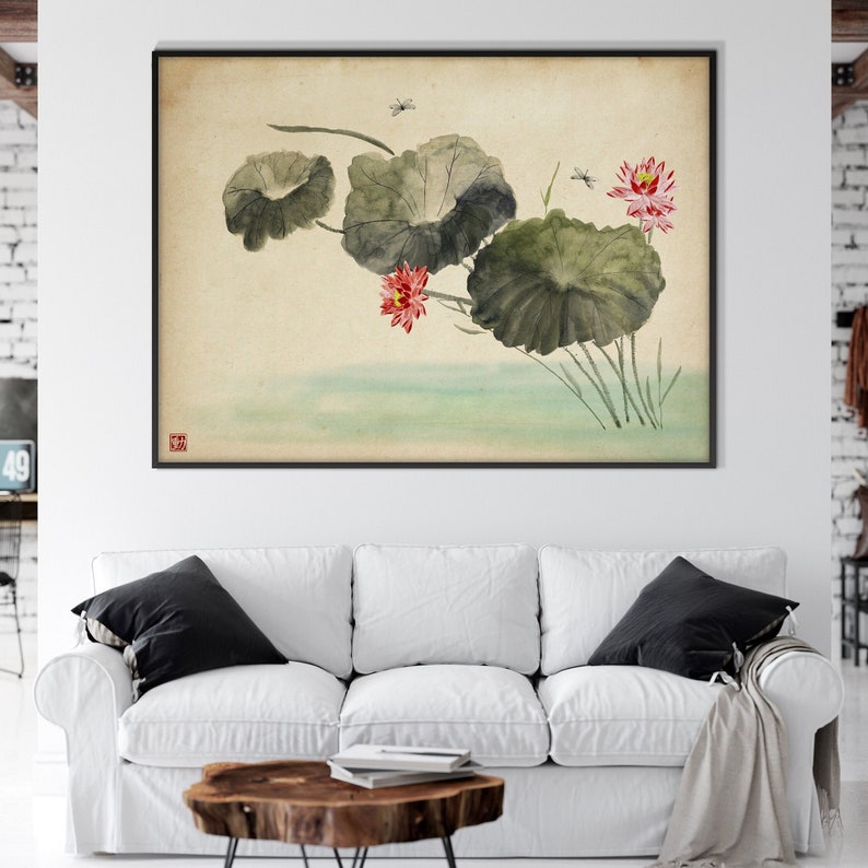 May include: A framed watercolor painting featuring lotus flowers and leaves in a traditional Asian style. The artwork showcases green leaves, pink flowers, and small insects against a beige background. The painting is displayed above a white sofa.