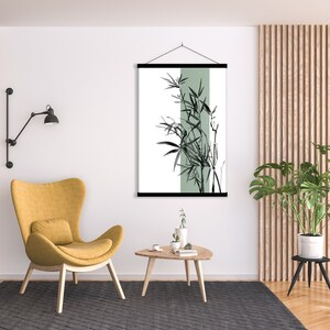 May include: A black and white illustration of bamboo plants with a green background hanging on a wall in a living room. The illustration is framed with a black wood frame and is hanging from a black cord. There is a yellow armchair, a wooden coffee table, and a potted plant in the room.