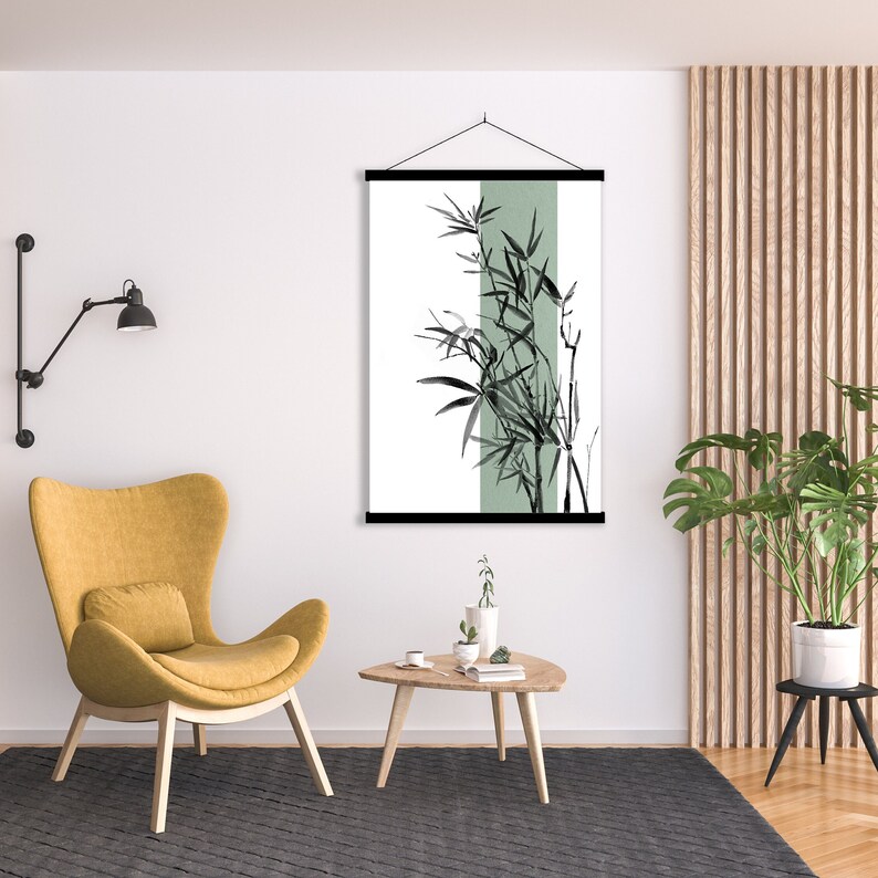 May include: A black and white illustration of bamboo plants with a green background hanging on a wall in a living room. The illustration is framed with a black wood frame and is hanging from a black cord. There is a yellow armchair, a wooden coffee table, and a potted plant in the room.