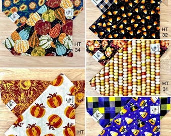 May include: Four different sets of dog bandanas featuring autumn and Halloween themes. The bandanas are made from fabric with various patterns, including pumpkins, sunflowers, candy corn, and ghosts. The bandanas are labelled with the text "HT" and a number.