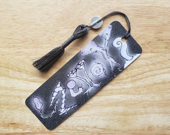 May include: A black bookmark with a gray tassel and a silver spiral charm. The bookmark features a hand-drawn design of mushrooms and snails on a gray background.