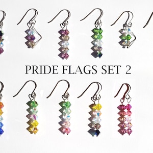 May include: Set of 10 pairs of pride flag earrings. Each pair features a different pride flag colour scheme with crystal beads. The earrings are silver and have a hook fastening. The text "PRIDE FLAGS SET 2" is printed below the earrings.