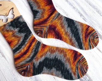 May include: A pair of hand-knitted socks in a colorful gradient pattern of orange, gray, and purple. The socks are displayed on wooden sock blockers.
