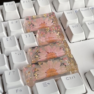 May include: Four custom keyboard keycaps with pink flowers and gold leaf pressed into clear resin. The keycaps are on a white keyboard.