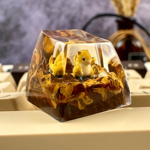 May include: A clear resin keycap with a brown and yellow floral design. A small yellow figurine of a creature is inside the keycap.
