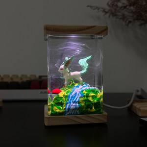May include: A wooden box with a clear glass insert containing a green and white figurine of a mythical creature. The figurine is standing on a green and blue base with white swirls. The box has a light inside that illuminates the figurine.