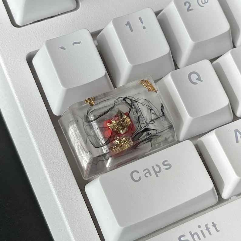 K&ouml;nnte beinhalten: Eine transparente Tastenkappe mit schwarzen Wirbeln und goldenen Flocken, darunter ein roter mechanischer Schalter. Die Tastenkappe befindet sich auf einer wei&szlig;en Tastatur mit dem Wort "Caps" auf der darunter liegenden Taste.