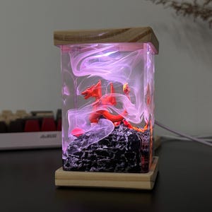 K&ouml;nnte beinhalten: Eine rote Drachenfigur, die in klarem Harz mit wirbelnden rosa und wei&szlig;en Rauchmustern eingeschlossen ist. Das Harz wird von innen beleuchtet und erzeugt ein sanftes rosa Leuchten. Der Drache sitzt auf einem schwarzen Felssockel.