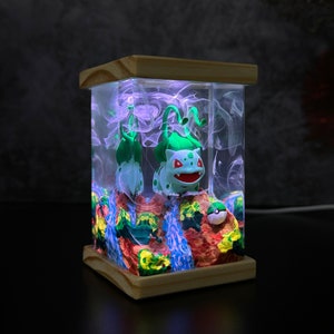 May include: A square resin art piece with a Bulbasaur figure and a Poke Ball, illuminated from within. The base and top are light-coloured wood. The interior features colourful, textured terrain and swirling, smoky effects.