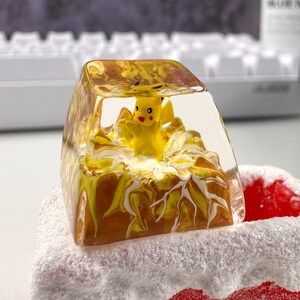 Może przedstawiać: Ż&oacute;łta figurka Pikachu jest zamknięta w przezroczystym, żywicznym klawiszu z wirującym brązowo-białym wzorem. Klawisz leży na biało-czerwonej powierzchni.