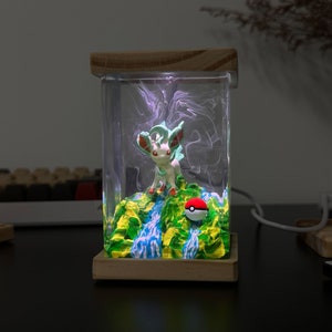 May include: A clear resin lamp with a wooden base features a small green and white figurine of a Pokemon character standing on a green and blue landscape with a red and white Pokeball. The lamp is lit with a white light.
