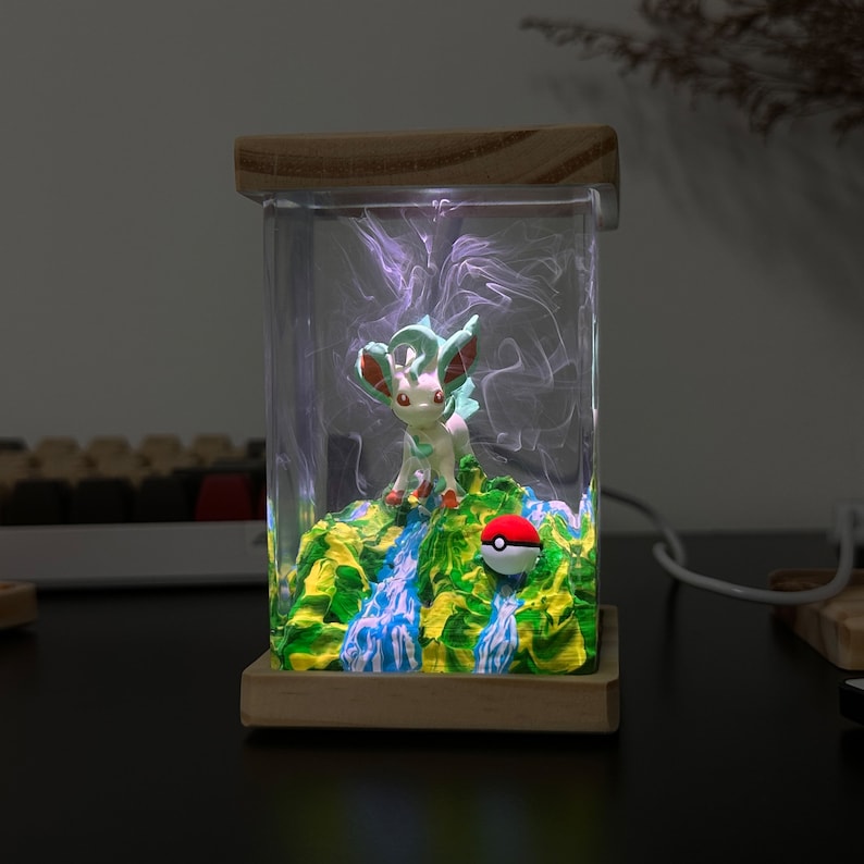 May include: A clear resin lamp with a wooden base features a small green and white figurine of a Pokemon character standing on a green and blue landscape with a red and white Pokeball. The lamp is lit with a white light.