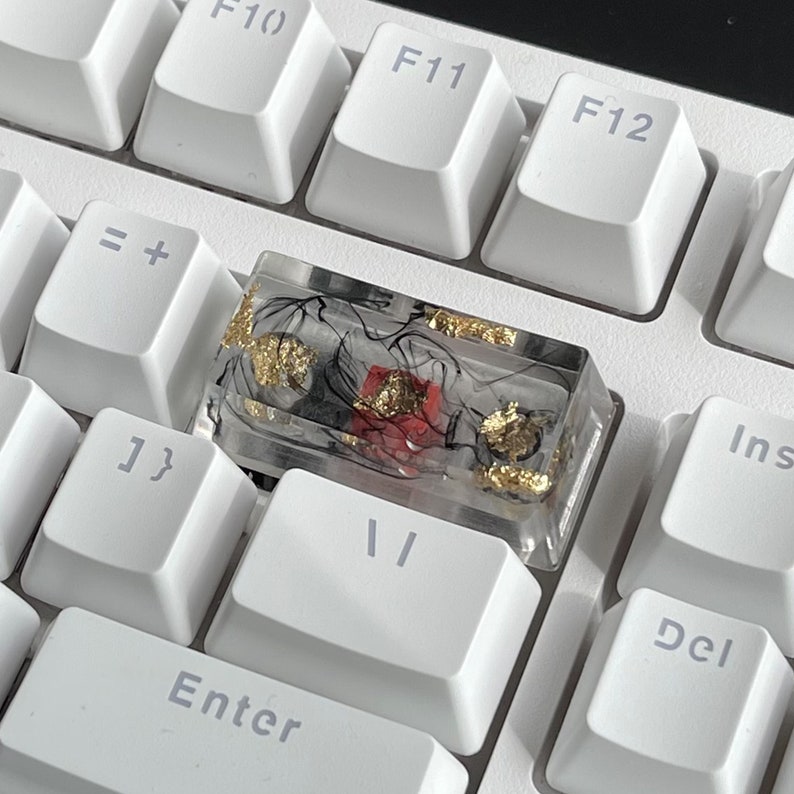 K&ouml;nnte beinhalten: Eine transparente Harztastenkappe mit schwarzen und goldenen Flocken und einem roten Akzent. Die Tastenkappe liegt auf einer wei&szlig;en Tastatur.