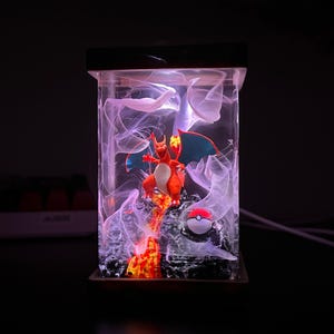 K&ouml;nnte beinhalten: Eine klare Harzlampe mit einer roten und orangefarbenen Pokemon-Figur, einem blauen und wei&szlig;en Pokeball und wei&szlig;en, rauch&auml;hnlichen Wirbeln im Inneren.
