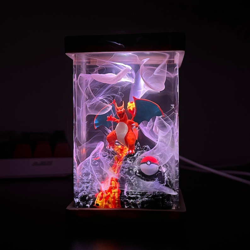 K&ouml;nnte beinhalten: Eine klare Harzlampe mit einer roten und orangefarbenen Pokemon-Figur, einem blauen und wei&szlig;en Pokeball und wei&szlig;en, rauch&auml;hnlichen Wirbeln im Inneren.