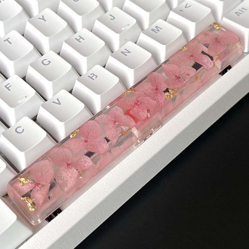 K&ouml;nnte beinhalten: Eine rosafarbene und goldene Tastaturkappe mit getrockneten Blumen, die in klarem Harz eingebettet sind. Die Tastaturkappe befindet sich auf einer wei&szlig;en Tastatur.