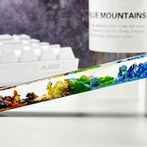 May include: A clear resin keycap with a colorful landscape scene inside. The scene features a blue mountain range, a green forest, a yellow field, and a brown rocky area. There are small figurines of Pokemon characters in the scene.