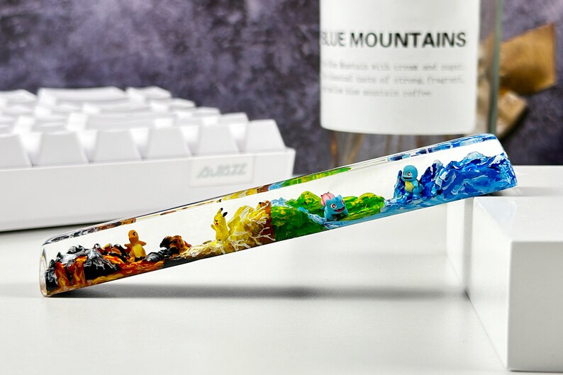 May include: A clear resin keycap with a colorful landscape scene inside. The scene features a blue mountain range, a green forest, a yellow field, and a brown rocky area. There are small figurines of Pokemon characters in the scene.