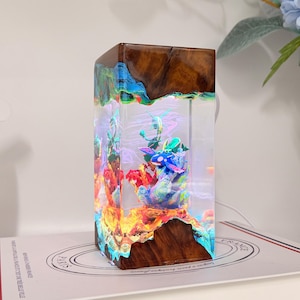 K&ouml;nnte beinhalten: Eine rechteckige Lampe aus dunkelbraunem Holz und klarem Kunstharz. Im Inneren des Harzes befindet sich eine farbenfrohe Unterwasserszene mit einem blau-wei&szlig;en Wesen inmitten von Korallen, das durch internes Licht durch das klare Material beleuchtet wird.