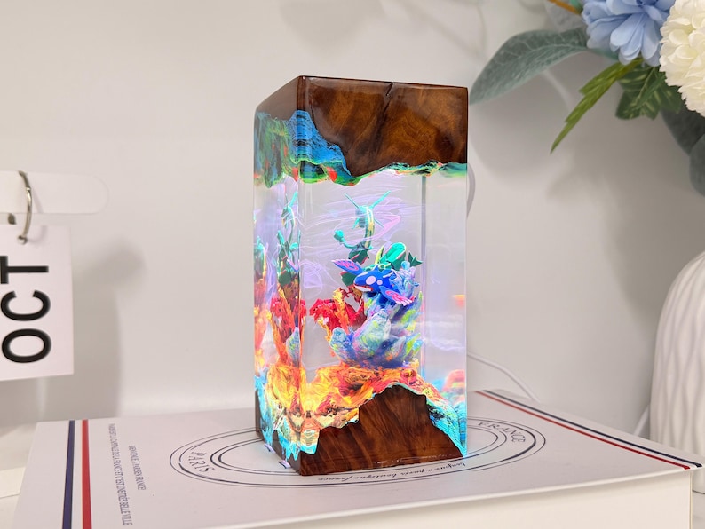 K&ouml;nnte beinhalten: Eine rechteckige Lampe aus dunkelbraunem Holz und klarem Kunstharz. Im Inneren des Harzes befindet sich eine farbenfrohe Unterwasserszene mit einem blau-wei&szlig;en Wesen inmitten von Korallen, das durch internes Licht durch das klare Material beleuchtet wird.