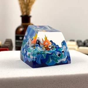 May include: A clear resin keycap with a blue, green, and white swirling design. Two orange and white fish figurines are inside the resin, swimming in the swirling design.