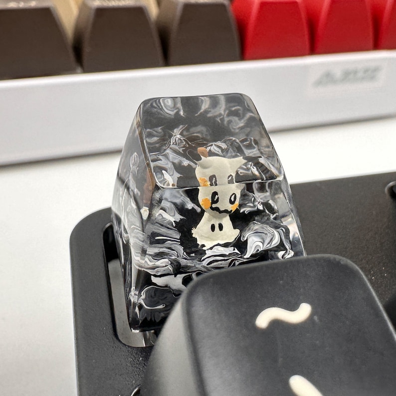 K&ouml;nnte beinhalten: Eine transparente, schwarz-wei&szlig;e marmorierte Tastenkappe mit einem kleinen wei&szlig;en und orangefarbenen Comic-Tier darin. Die Tastenkappe befindet sich auf einer schwarzen Tastatur.