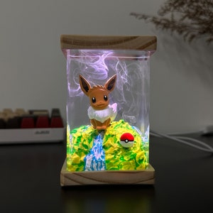 May include: A clear glass lamp with a wooden base features a brown and white Eevee figurine standing on a green, yellow, and blue landscape. A red and white Pokeball sits on the landscape. The lamp is lit with a purple glow.