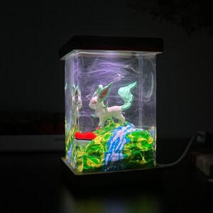 May include: A clear glass lamp with a wooden base features a green and blue resin landscape with a white Eevee figurine. The lamp is lit and casts a soft glow.