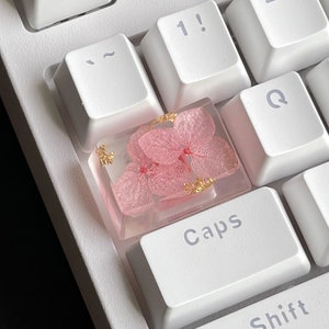 May include: A pink and gold resin keycap with dried flowers embedded inside. The keycap is labeled "Caps" and is sitting on a white keyboard.
